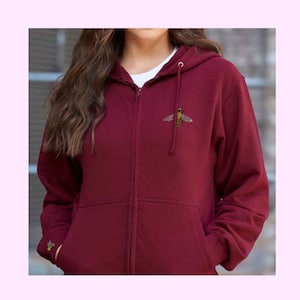 May include: A maroon zip-up hoodie with a small embroidered bee on the front. The hoodie has a hood with a drawstring and two front pockets.