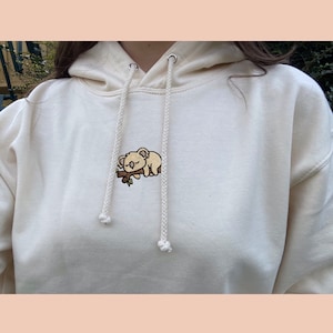 May include: A cream-coloured hoodie with a small embroidered koala sleeping on a branch. The hoodie has a drawstring hood and a relaxed fit.