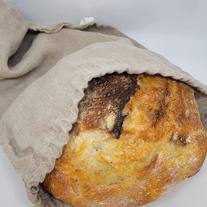 May include: A loaf of freshly baked sourdough bread in a linen bread bag. The bread has a dark brown crust and a light brown interior.