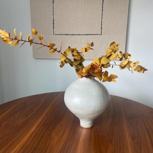 Puede incluir: Un jarrón de cerámica blanco y redondo con una base estrecha contiene ramas con hojas amarillas y marrones. El jarrón está sobre una mesa de madera oscura. Un lienzo de tonos neutros con un diseño cuadrado negro cuelga en el fondo.