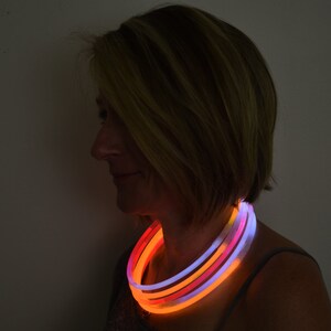 May include: A multi-layered glow necklace featuring flexible, illuminated tubes in vibrant shades of orange, pink, and white. The circular light-up accessory rests around the neck, emitting a soft, radiant glow against a dark background.