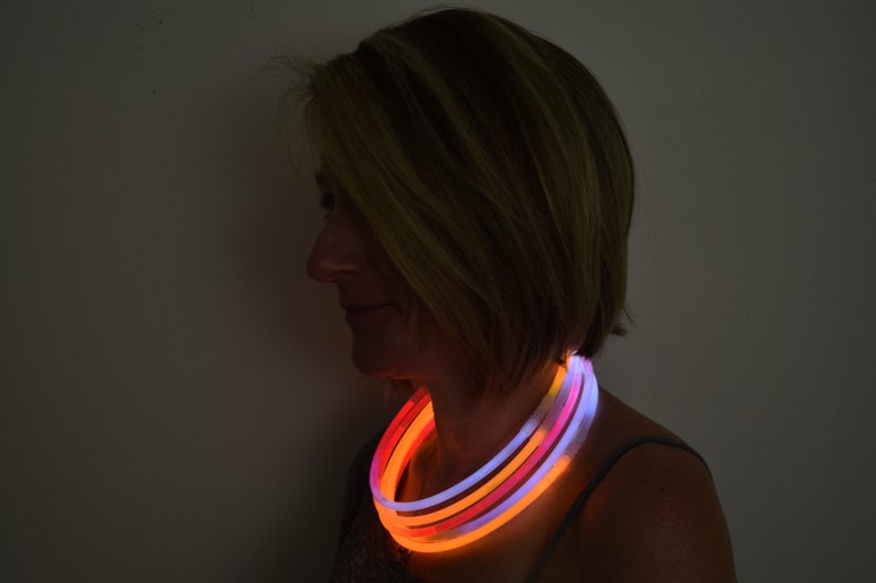 May include: A multi-layered glow necklace featuring flexible, illuminated tubes in vibrant shades of orange, pink, and white. The circular light-up accessory rests around the neck, emitting a soft, radiant glow against a dark background.