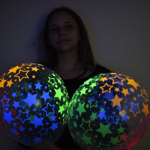 May include: Two glow-in-the-dark balloons with a star pattern in shades of blue, green, orange, and yellow. The balloons are held by a person in a dark room.