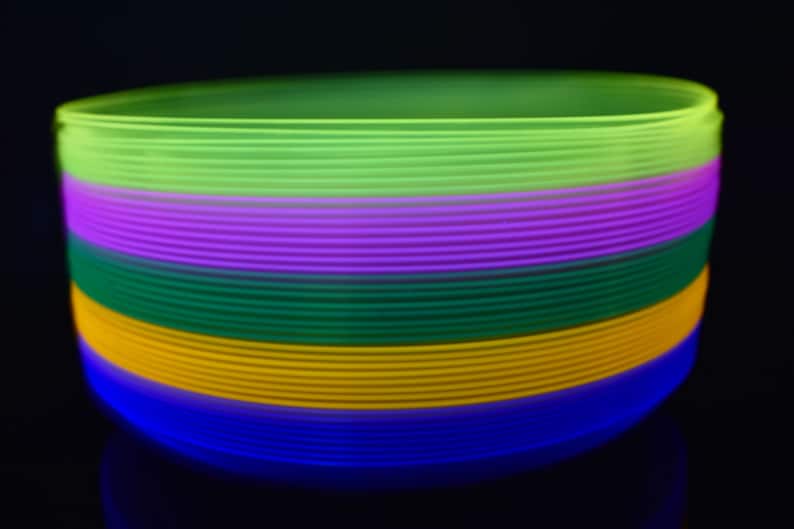 May include: A stack of colorful glow sticks in green, purple, blue, yellow, and orange. The glow sticks are stacked on top of each other and are glowing in the dark.