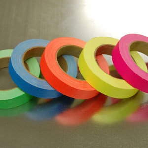 K&ouml;nnte beinhalten: F&uuml;nf Rollen farbiges Klebeband, darunter gr&uuml;n, blau, orange, gelb und pink. Das Klebeband ist um Pappkerne gewickelt.