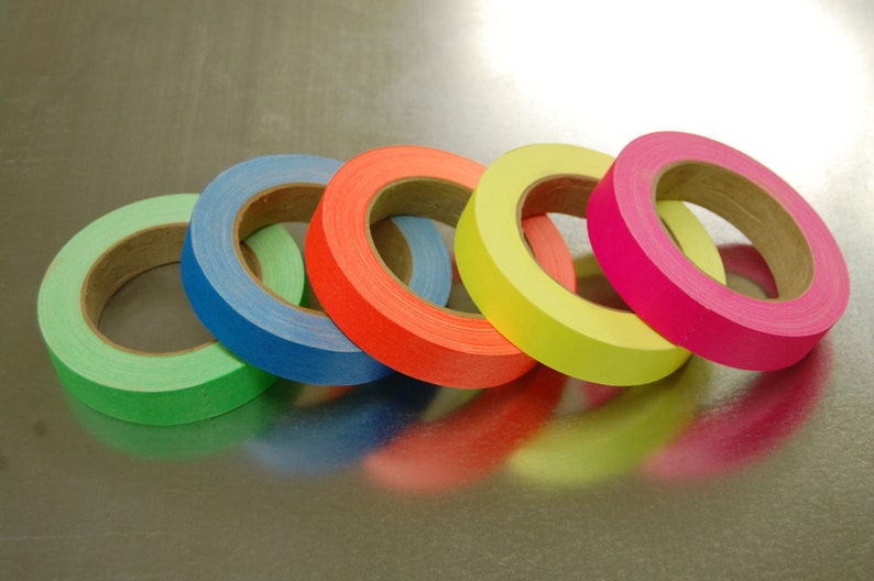 K&ouml;nnte beinhalten: F&uuml;nf Rollen farbiges Klebeband, darunter gr&uuml;n, blau, orange, gelb und pink. Das Klebeband ist um Pappkerne gewickelt.