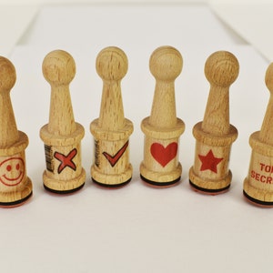 May include: Set of seven wooden stamps with different designs. The designs include a smiley face, a red X, a red checkmark, a red heart, a red star, and the words "Top Secret".