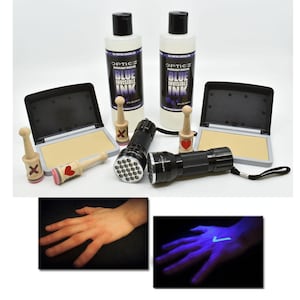 May include: An assortment of items including two bottles of blue invisible ink, two black flashlights, two ink pads, and several wooden stamps with red heart and X designs. The image also shows a hand with a mark that glows under UV light.