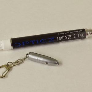 May include: A black marker labeled "OPTICZ INVISIBLE INK" with a white tip and a small silver keychain light. The text "Premium Quality. Glows bright BLUE" is also visible on the marker. The light is attached to a silver keychain.