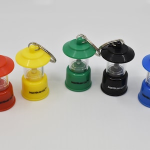 May include: Five miniature lantern keychains in red, yellow, green, black, and blue. Each lantern features a clear plastic center, a solid colored base and top, and a metal split ring. The text DirectGlow LLC is printed on each base.