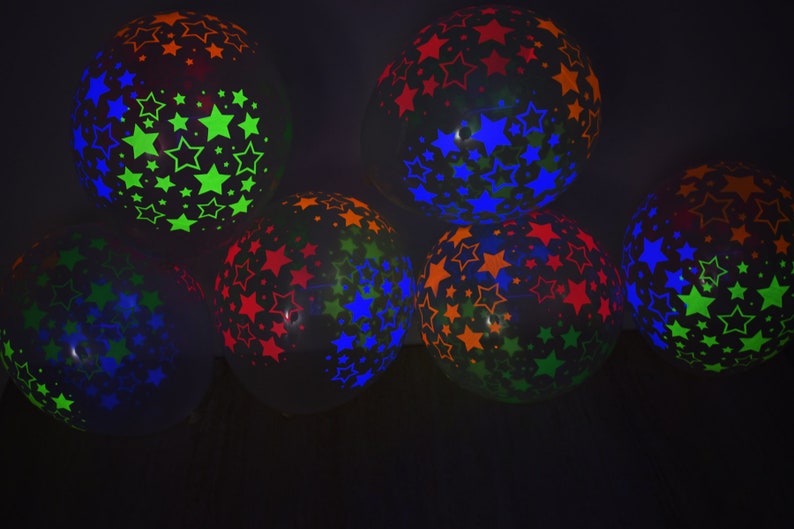 May include: A group of six black balloons with neon star patterns. The balloons are filled with helium and are floating in the air. The stars are in different colors, including green, blue, orange, and red.