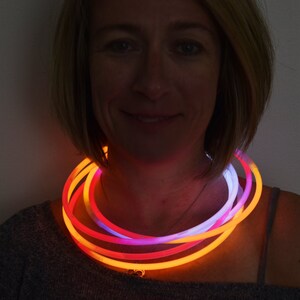 May include: A glowing necklace composed of multiple flexible tubes that emit bright orange, pink, and white light. The circular tubes are layered around the neck, creating a vibrant, multi-colored illumination effect that stands out against the dark.