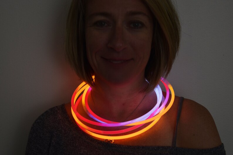 May include: A glowing necklace composed of multiple flexible tubes that emit bright orange, pink, and white light. The circular tubes are layered around the neck, creating a vibrant, multi-colored illumination effect that stands out against the dark.