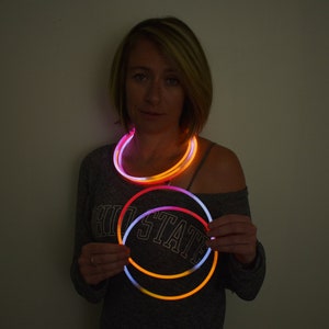 May include: A set of circular glow necklaces emitting bright orange and white light. The flexible, tubular accessories are illuminated, creating a vibrant contrast against the dark environment. These wearable light-up items are designed to be worn around the neck or held in the hands as glowing rings.
