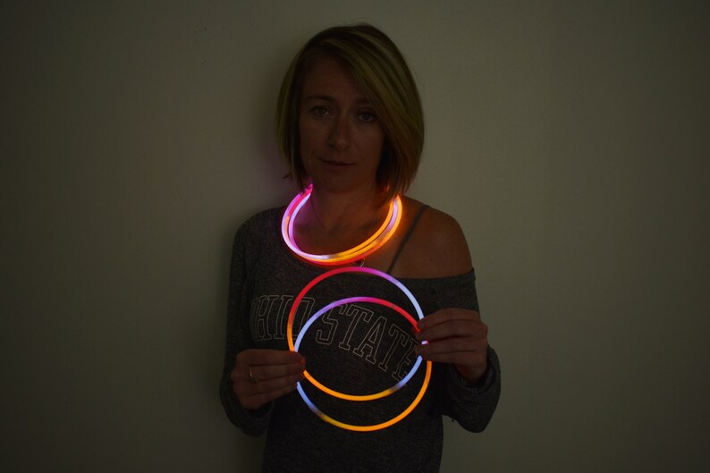 May include: A set of circular glow necklaces emitting bright orange and white light. The flexible, tubular accessories are illuminated, creating a vibrant contrast against the dark environment. These wearable light-up items are designed to be worn around the neck or held in the hands as glowing rings.