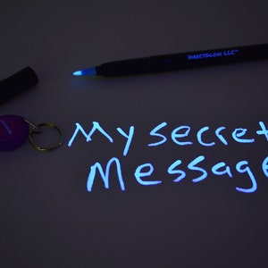 May include: A black UV pen and a purple UV light keychain are shown on a dark surface. The words "My Secret Message" are written in white ink that is only visible under the UV light.