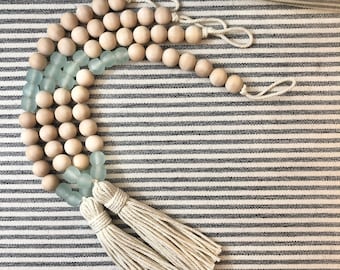 May include: A wooden bead garland with light blue glass beads and two white tassels. The garland is on a striped fabric background.