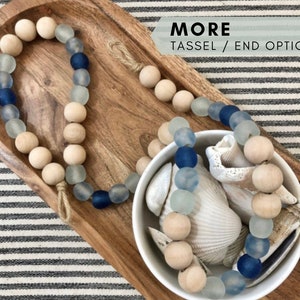 May include: A wooden and glass bead garland with blue and white beads. The garland is displayed on a wooden tray and in a white bowl with seashells. The text "MORE TASSEL / END OPTIONS" is displayed in the top right corner of the image.