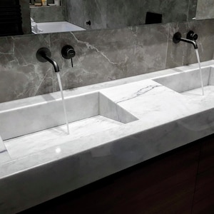 Peut inclure: Un meuble-vasque double en marbre blanc avec deux robinets noirs, dont l'un est en marche. Le meuble-vasque est monté sur un meuble en bois foncé.