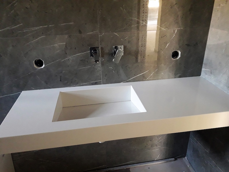 Puede incluir: Un lavabo de ba&ntilde;o de encimera blanco con una cuenca rectangular. El lavabo est&aacute; montado en una pared con azulejos de m&aacute;rmol gris.