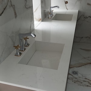 Puede incluir: Dos lavabos de ba&ntilde;o de porcelana blanca con grifos cromados y detalles dorados. Los lavabos est&aacute;n montados en una encimera marr&oacute;n con un salpicadero de m&aacute;rmol blanco.