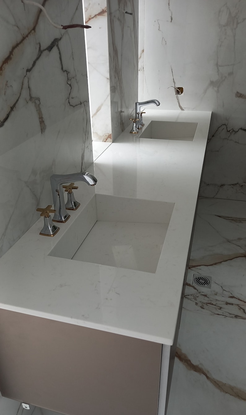 Puede incluir: Dos lavabos de ba&ntilde;o de porcelana blanca con grifos cromados y detalles dorados. Los lavabos est&aacute;n montados en una encimera marr&oacute;n con un salpicadero de m&aacute;rmol blanco.