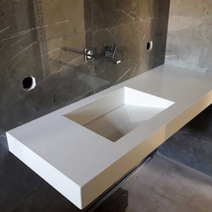 Puede incluir: Una encimera blanca con un lavabo rectangular recortado. La encimera est&aacute; montada en una pared con azulejos de m&aacute;rmol gris.