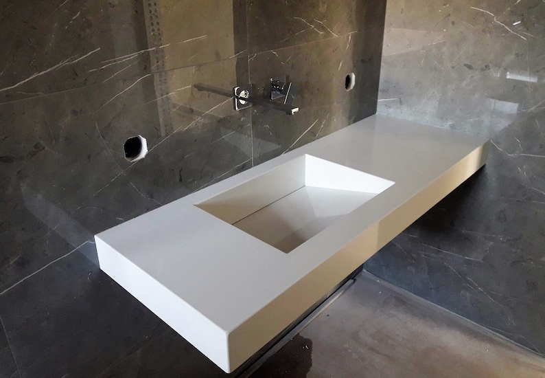 Puede incluir: Una encimera blanca con un lavabo rectangular recortado. La encimera est&aacute; montada en una pared con azulejos de m&aacute;rmol gris.