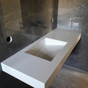 Puede incluir: Un lavabo de ba&ntilde;o de encimera blanco con un recorte rectangular para la cuenca. El lavabo est&aacute; montado en una pared con azulejos de m&aacute;rmol gris.