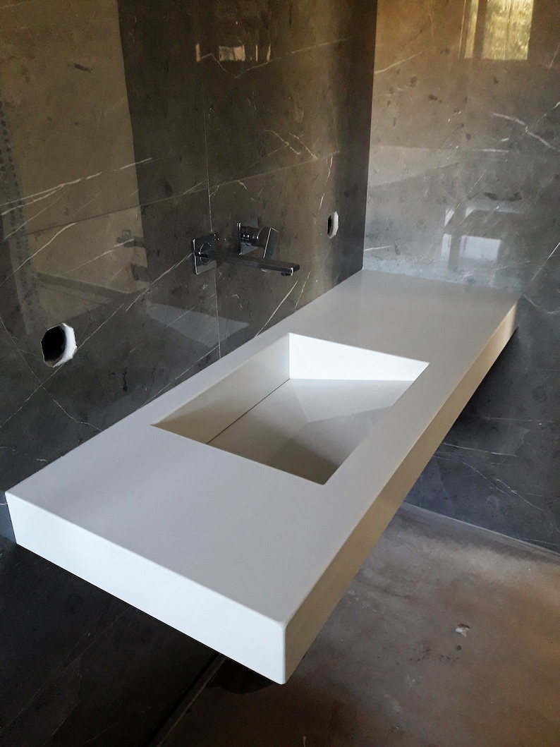 Puede incluir: Un lavabo de ba&ntilde;o de encimera blanco con un recorte rectangular para la cuenca. El lavabo est&aacute; montado en una pared con azulejos de m&aacute;rmol gris.