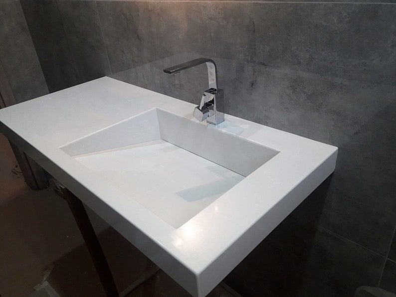Puede incluir: Un lavabo de ba&ntilde;o de encimera blanco con un grifo cromado. El lavabo es rectangular con un borde ligeramente inclinado.