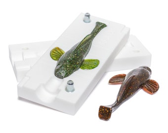 May include: A white plastic mold for making fishing lures, with two finished lures, one green and one brown, sitting next to it.
