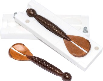 May include: A white plastic mold for making fishing lures. The mold is open and shows a brown lure with a split tail. The lure is made of soft plastic and has a ribbed body.