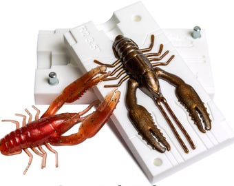May include: A red and brown crawfish lure next to a white mold with the text "R2 3.5" on it. The crawfish lure is a detailed replica, with visible claws, legs, and antennae. The mold is open, showing the crawfish shape.