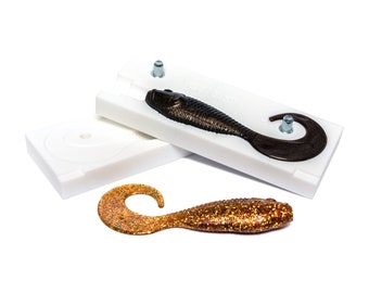 May include: Two white plastic molds for making fishing lures, one with a dark brown lure inside, and a separate gold lure with glitter.