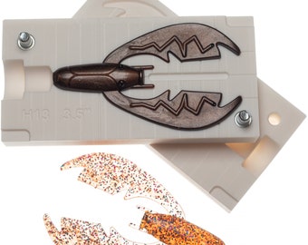 May include: A white plastic mold for making fishing lures. The mold is open and shows a brown lure with a textured surface. The mold is labeled "H13 3.5". A finished lure is shown below the mold. The lure is translucent with a brown body and a glittery finish.