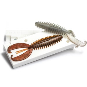 以下が含まれることがあります： 白いプラスチック製の型の中に、茶色と灰色のプラスチック製の釣りルアーが2つあります。ルアーはセグメント化されたボディとフォーク状のテールを持っています。