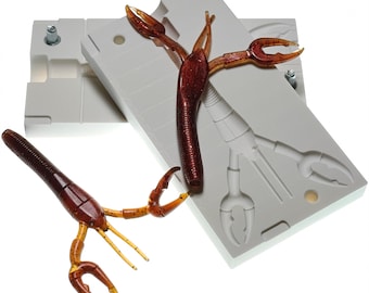 May include: Two brown and orange crawfish lures with a gray mold. The lures have detailed claws and antennae. The mold has a detailed crawfish impression. The background is white.