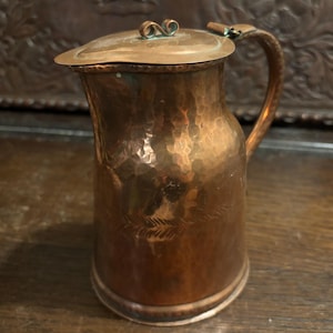 May include: A hammered copper jug with a hinged lid and a curved handle. The metal surface features a warm, reddish-brown patina with visible oxidation and a subtle engraved leaf-like pattern encircling the lower body of the vessel.