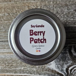 Puede incluir: Una vela en una lata plateada con una etiqueta blanca que dice "Soy Candle Berry Patch" y "Clohi's Candle Corner" con iconos de redes sociales.