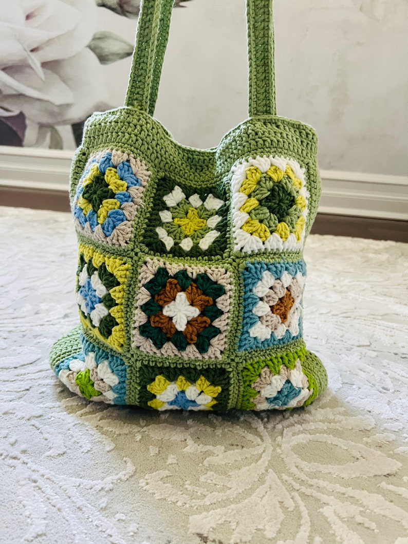 K&ouml;nnte beinhalten: Eine gr&uuml;ne geh&auml;kelte Tragetasche mit Patchwork-Muster aus bunten Quadraten. Die Quadrate sind in den Farben Gr&uuml;n, Gelb, Blau, Wei&szlig; und Braun gehalten.