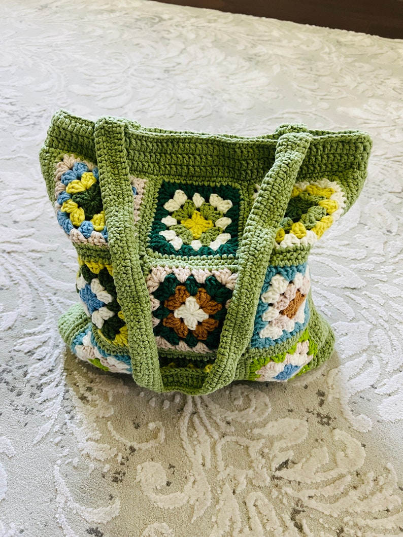 K&ouml;nnte beinhalten: Eine gr&uuml;ne geh&auml;kelte Tragetasche mit bunten quadratischen Mustern. Die Tasche hat zwei Henkel und ist mit einem Granny-Square-Stich gearbeitet.
