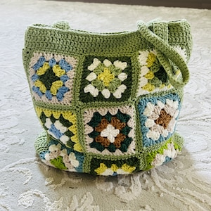 K&ouml;nnte beinhalten: Gr&uuml;ne geh&auml;kelte Tragetasche mit einem bunten Granny-Square-Muster. Die Tasche hat zwei Henkel und ist aus verschiedenen gr&uuml;nen, wei&szlig;en, braunen, blauen und gelben Garnen gefertigt.