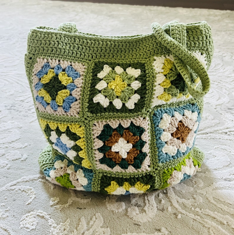 K&ouml;nnte beinhalten: Gr&uuml;ne geh&auml;kelte Tragetasche mit einem bunten Granny-Square-Muster. Die Tasche hat zwei Henkel und ist aus verschiedenen gr&uuml;nen, wei&szlig;en, braunen, blauen und gelben Garnen gefertigt.