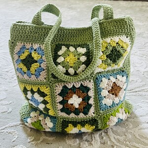 K&ouml;nnte beinhalten: Eine gr&uuml;ne geh&auml;kelte Tragetasche mit Patchwork-Muster aus bunten Quadraten. Die Tasche hat zwei lange Henkel und ist mit einem Granny-Square-Stich gearbeitet.
