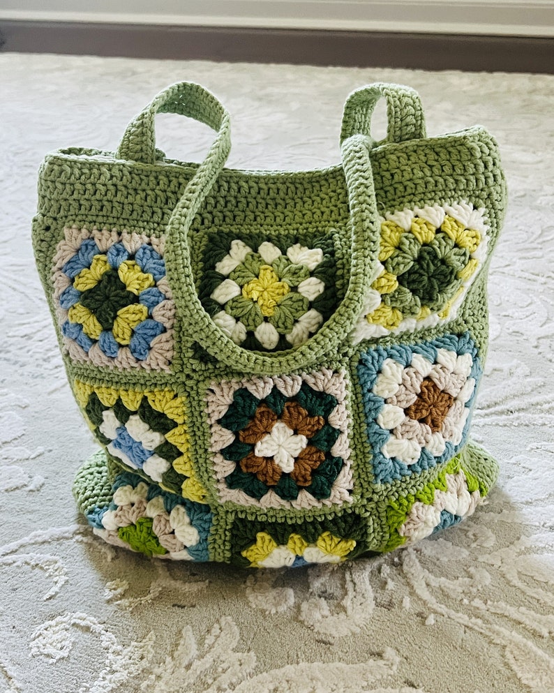 K&ouml;nnte beinhalten: Eine gr&uuml;ne geh&auml;kelte Tragetasche mit Patchwork-Muster aus bunten Quadraten. Die Tasche hat zwei lange Henkel und ist mit einem Granny-Square-Stich gearbeitet.