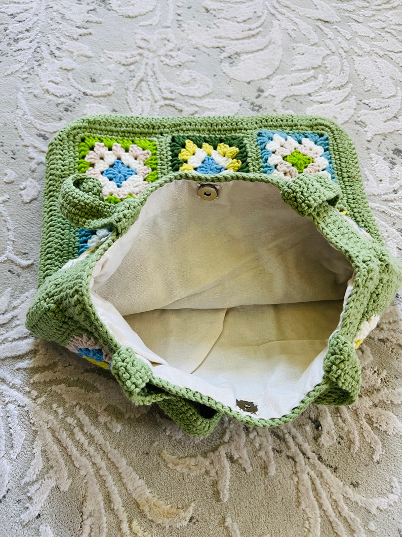 K&ouml;nnte beinhalten: Gr&uuml;ne geh&auml;kelte Tragetasche mit Blumenmuster. Die Tasche ist offen und das Innere ist mit einem wei&szlig;en Stoff ausgekleidet.