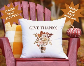 May include: White throw pillow with the text "GIVE THANKS" printed in brown. The pillow features a watercolor illustration of a mason jar filled with fall leaves, flowers, and berries.
