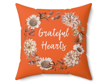 May include: Orange throw pillow with a floral wreath design in shades of brown and white. The wreath has the text "Grateful Hearts" in white.