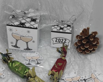 May include: Two small gift boxes with silver bows, one with champagne glasses and the other with the year 2022. Also, two wrapped candies and a pine cone are on a white surface. The boxes have a black and white plaid pattern.